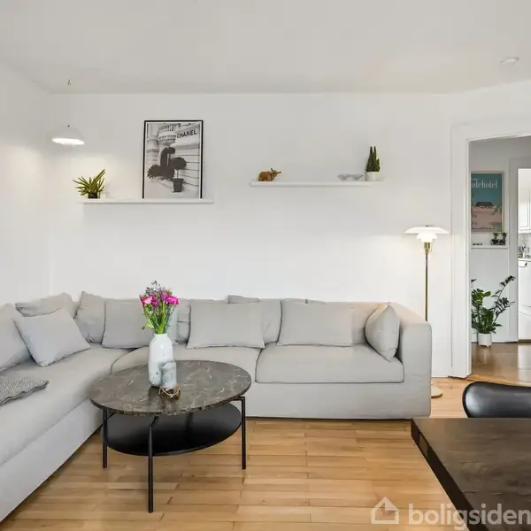 En grå hjørnesofa står i en lys stue. På sofabordet er en vase med blomster, og væggen har dekorative hylder. Rummet fører til et køkken.