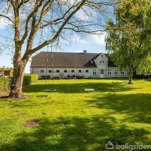 Et stort, gammelt hus med sort tag står omgivet af en frodig have. Gynger hænger fra et stort træ, og græsplænen er omkranset af buske og andre træer.