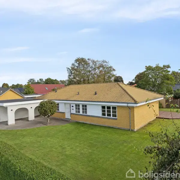 Gult murstenshus med gul tagstenstag står i en velplejet have med græsplæne og en hæk. Til højre for huset ses en garage. Omkring er der andre boliger og træer.