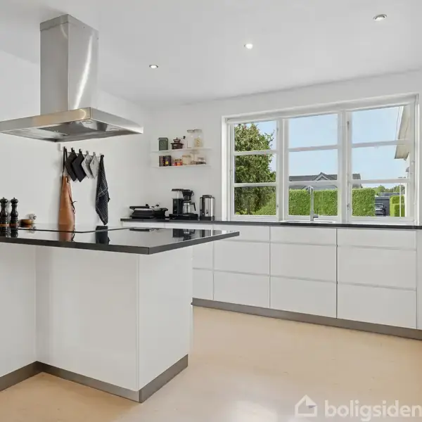 Et moderne køkken med hvide skabe og en sort bordplade. Emhætten hænger over en ø. Vinduet viser en have i baggrunden. Krydderier og køkkenredskaber ses på bordet.