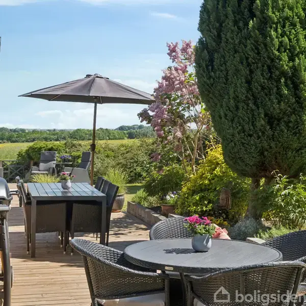 Grill står på træterrasse, omgivet af havemøbler under parasol. Lige ved siden af står et stort, grønt cypreslignende træ i en frodig have med udsigt til marker.