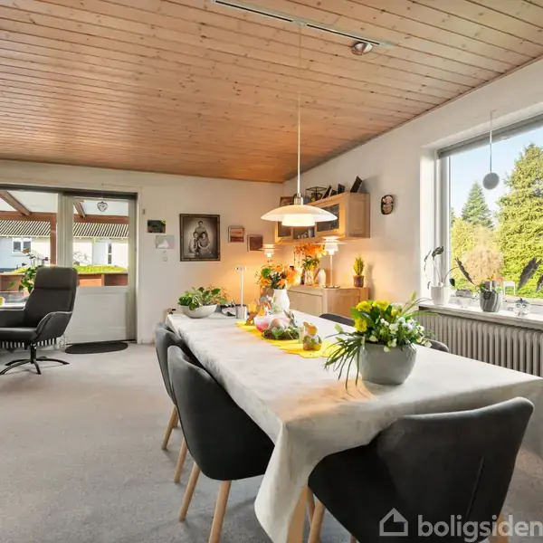 Et spisebord fyldt med blomsterdekorationer står i en spisestue. Rummet har lyse vægge og træloft, med store vinduer der giver udsigt til en have med træer.