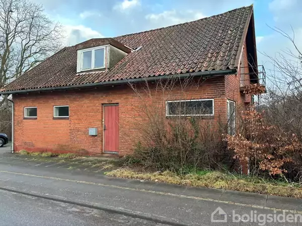 Rødt murstenshus med rødt tag ser gammelt og slidt ud, står langs en vej med overgroede buske omkring. Huset har en enkelt dør og flere rektangulære vinduer.