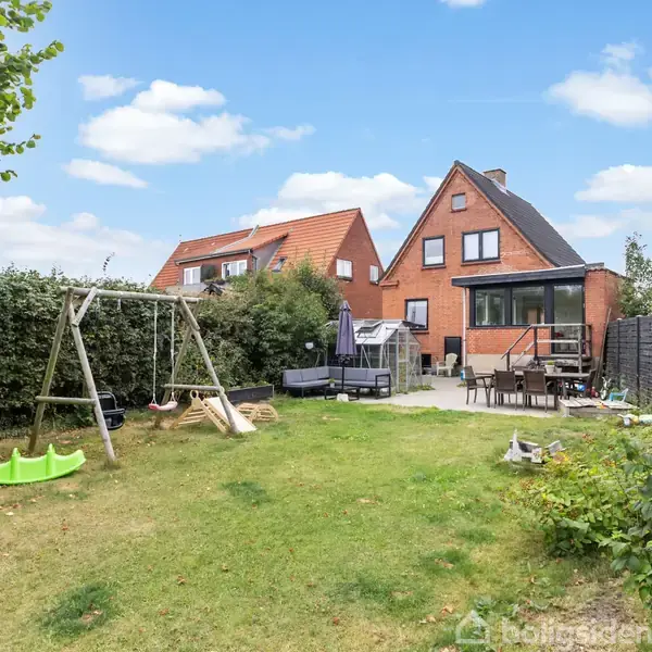 Et gyngestativ står på en græsplæne i en baghave med en hækomgivet terrasse. I baggrunden ses et rødt murstenshus med skråt tag og hvide vinduer.