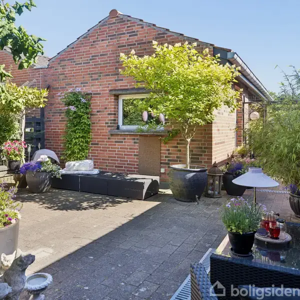 Rød murstensgårdhave med plantekrukker og havemøbler. Et lille træ og grønne planter langs væggen. Klar himmel over gården; afslappet atmosfære med komfortable siddepladser og dekorationer.