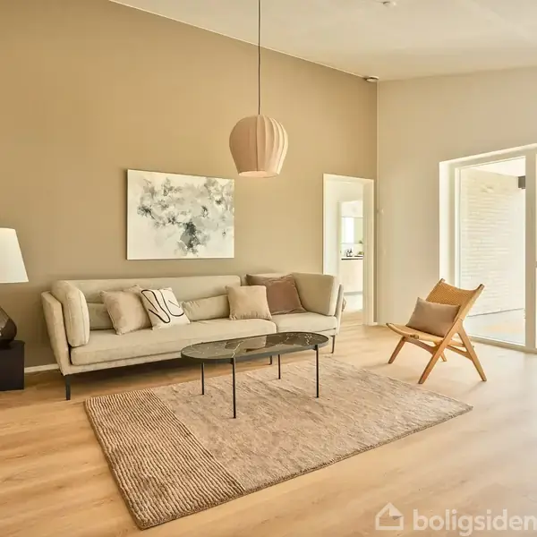 Sofa med puder står i stue, omgivet af bord, stol og lampe. På væggen hænger kunstprint. Rummet er lyst med store vinduer og en åben dør til altan.