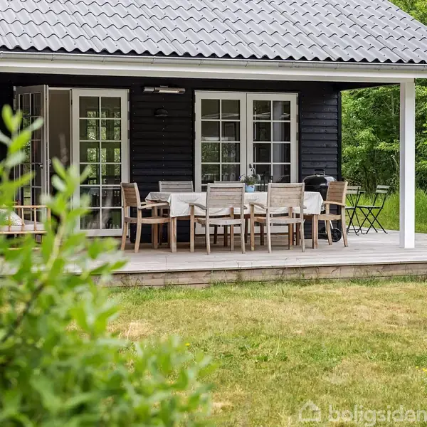 En terrasse med et bord og flere træstole står foran et sort hus med hvide vinduer. Omkring er der en grøn græsplæne og buske.