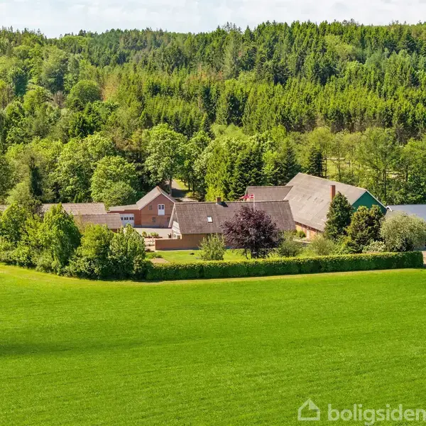 En gård med flere bygninger ligger omgivet af frodige grønne marker og en tæt skov i baggrunden. Det hele skaber en idyllisk, landlig atmosfære.