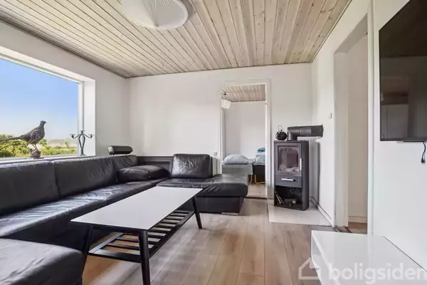 Sort hjørnesofa står i en moderne stue, mens et kaffebord er placeret foran. Et stort vindue giver udsigt til træer. Væggene er hvide, og der er trægulve.