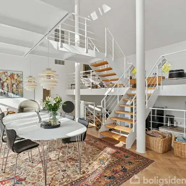 Rundt bord med sorte stole står på et dekorativt tæppe. Trappe fører til øvre niveau med sofaer. Store vinduer og moderne kunst præger den åbne, lyse stue.