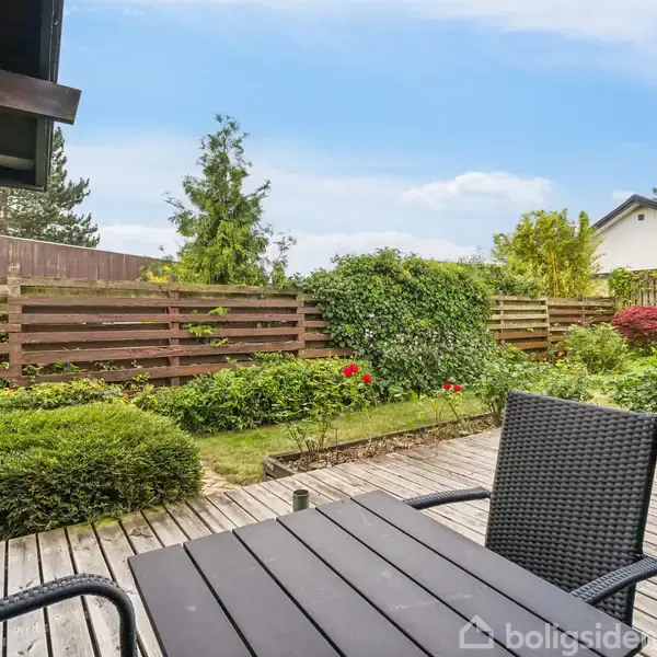 En terrassebord med to stole står på en træterrasse. Haven er omgivet af et lavt hegn med buske og grønt nær et hus i baggrunden.