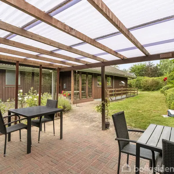 To borde med stole står under en overdækket terrasse. Omkring dem er der en flisebelagt gårdhave og en have med græsplæne, træer og en træbygning i baggrunden.