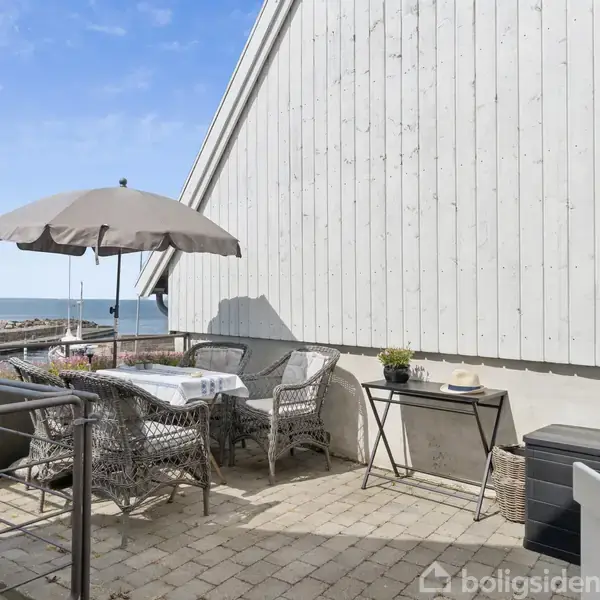 Terrasse med bord og stole under parasol. Omgivet af blomsterkrukker, står møblerne ved siden af et hvidt træhus med udsigt over havet.