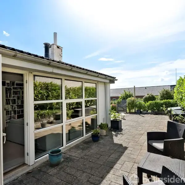 En terrasse med fliser har sorte havemøbler og krukker. Store vinduer i baggrunden leder ind til et hus, omgivet af grønne buske og en klar blå himmel.