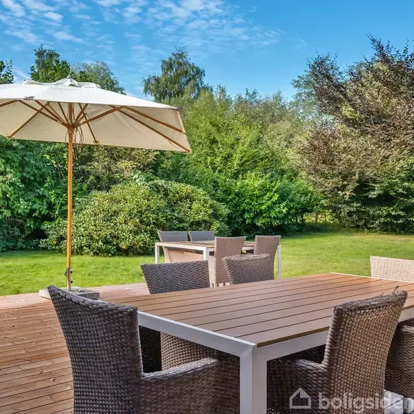 En terrasse med et bord og seks kurvestole står under en stor parasol. Baggrunden viser en grøn have med træer og buske.