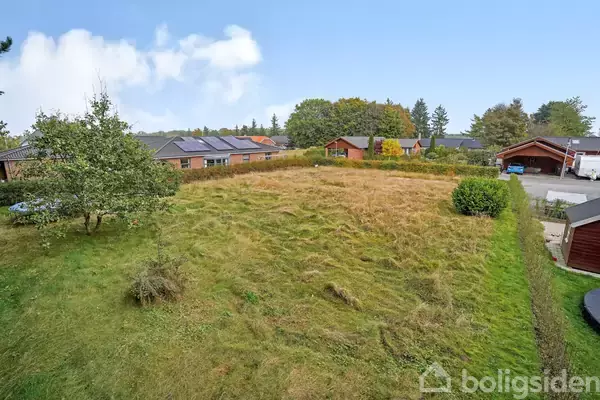 En åben græsmark med en enkelt træ, der står i nærheden af en række huse med solpaneler på taget. Området er omgivet af større træer i baggrunden.