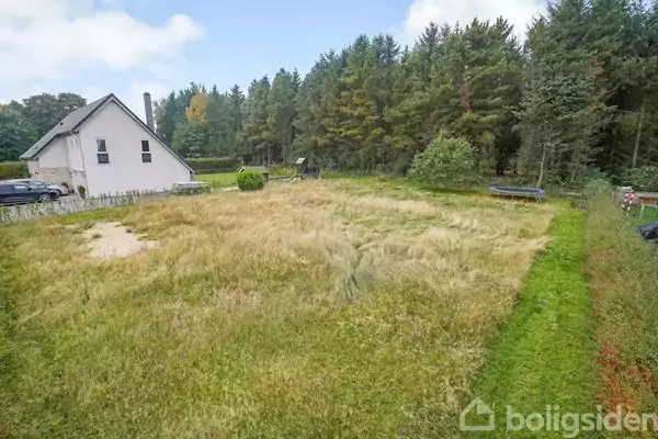 En hvid murstensvilla står ved siden af en stor, åben græsplæne omgivet af et tætte træer, med en lille terrasse og opbevaringsbænk. Biler er parkeret ved huset.