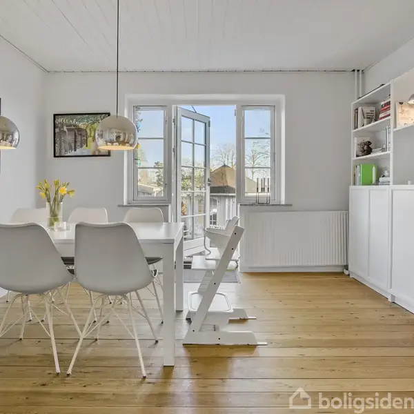 Spisebord med hvide stole står i en lys spisestue. To hængelamper hænger over bordet. Væghylde og åben terrassedør viser udsigt til en have. Tulipaner på bordet.