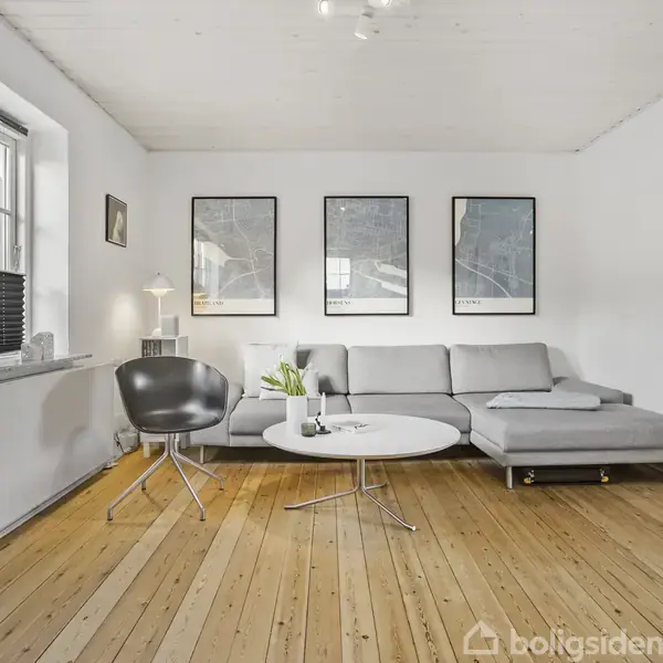 Grå sofa står i ro i en lys stue med trægulv, flankeret af en sort stol og hvidt rundt sofabord. Væggene prydes af tre indrammede billeder.