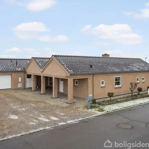 En moderne, enfamilies murstensvilla med mørkt tegltag står på en flisebelagt indkørsel. Til venstre er en garage, og hele huset er omgivet af landligt landskab under en skyet himmel.