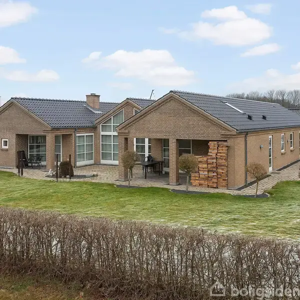 En murstensvilla med moderne arkitektur står på en velplejet græsplæne, omkranset af en hæk. Villaen har store vinduer og en terrasse med havemøbler.