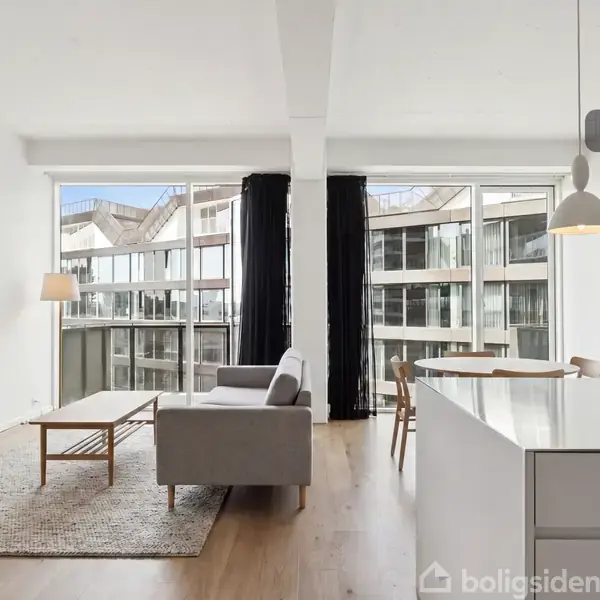 En moderne stue med en grå sofa og træbord placeret på et tæppe. Ved siden af er et køkkenområde med spisebord. Store vinduer åbner mod en bygning udenfor. "Visualisering" vises øverst.