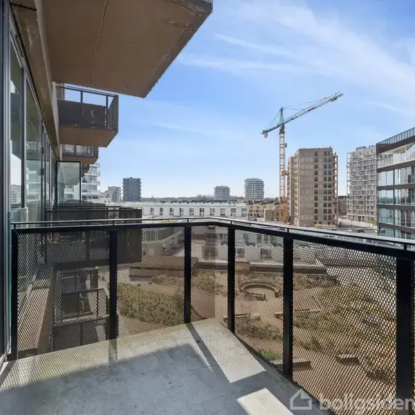 Balkon set fra en moderne lejlighedskompleks, vendt mod en byudsigt med flere konstruktioner og en kran i baggrunden under en klar himmel.