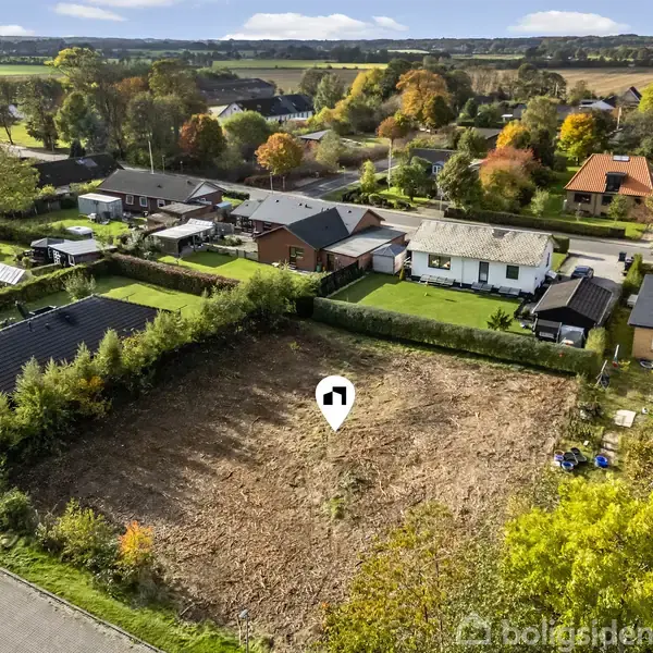 En tom byggegrund markeret med en hvid nål ligger i et boligområde omgivet af rækkehuse og grønne haver, med en vej foran og landlige marker i baggrunden.
