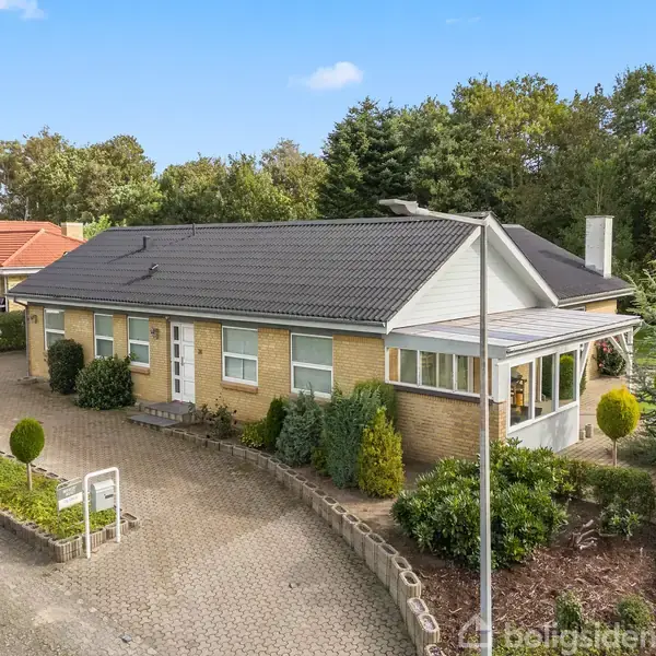 En etplanshus med sort tag står stille omgivet af en flisebelagt indkørsel og veltrimmet have i et villakvarter. Træer og græs danner en grøn baggrund.