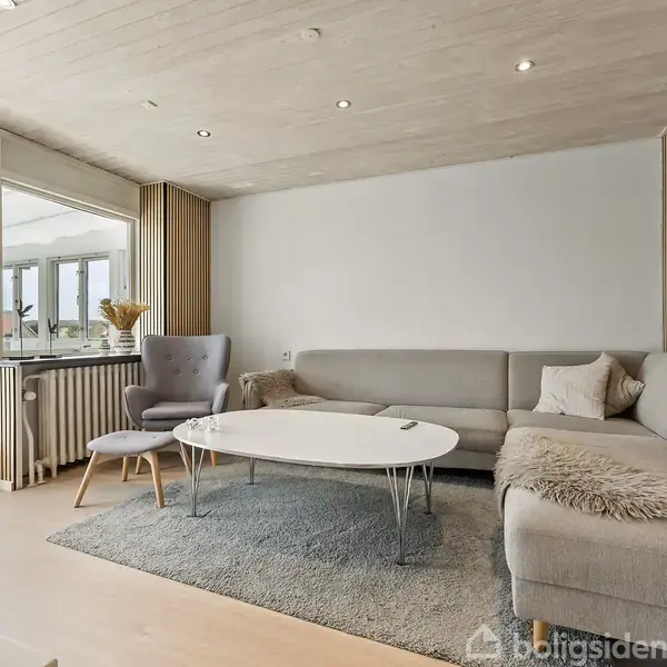 Grå sofa står i hjørne, omgivet af et sofabord og lænestol på gråt tæppe, i en stue med trægulv og store vinduer, der åbner til en glasveranda.