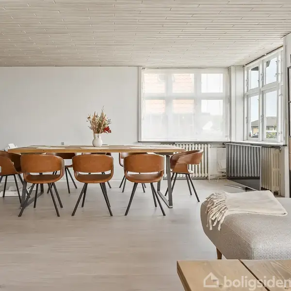 Brændeovn står ved væggen og opvarmer spisestue med træbord og otte stole. Store vinduer lukker lys ind, reol står til højre, sofa i forgrunden.