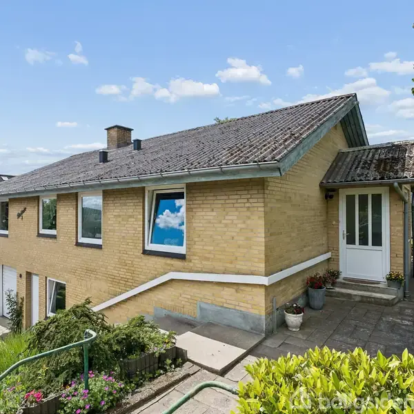 Et gult murstenshus med store vinduer står på en skrånende grund. Foran huset er der en trappe, og omgivelserne inkluderer en have med buske og træer under en blå himmel.