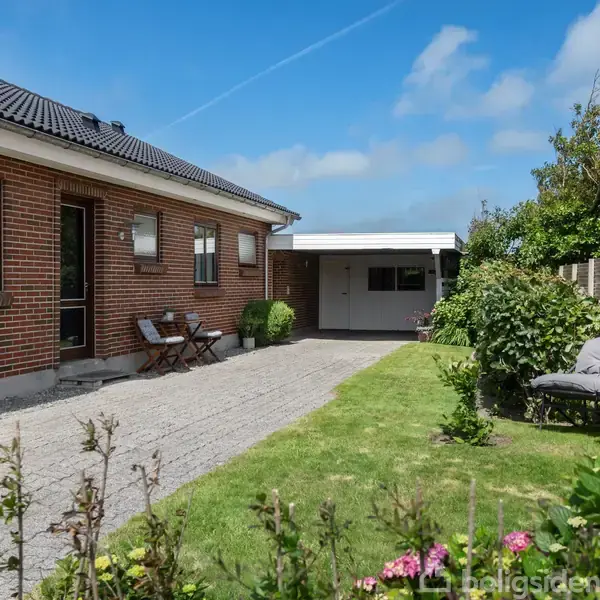 Et murstenshus med sorte tagsten står ved en grussti. To havestole står ved siden af huset, og en grøn have med buske og blomster omgiver området under en blå himmel.