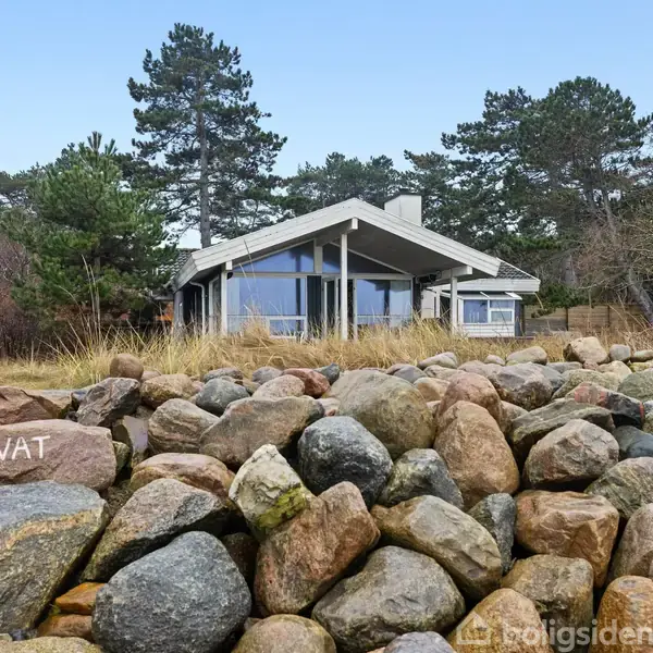 Hus står stationært bag en stenmuret have med store, runde klippesten. Omkringliggende natur består af træer og buskads. Tekst på sten: "PRIVAT".