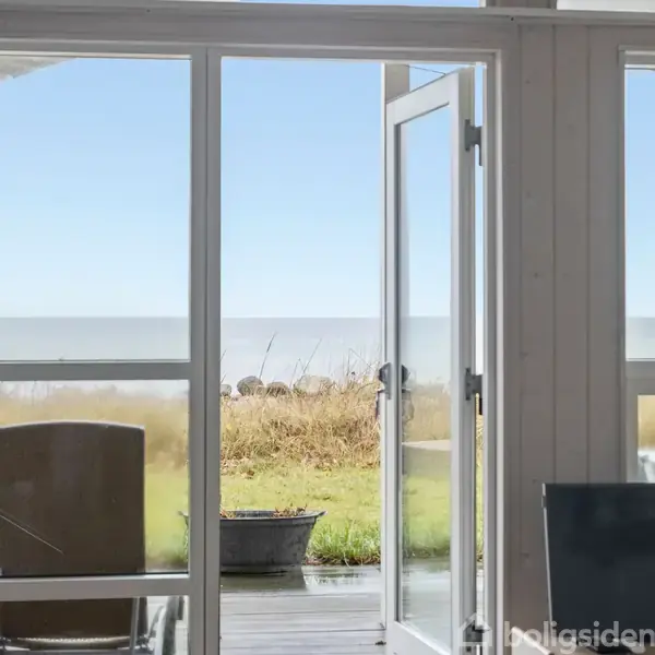 En glasdør står åben, hvilket giver udsigt til en terrasse med havemøbler. I baggrunden ses græsarealer og havet under en klar himmel.
