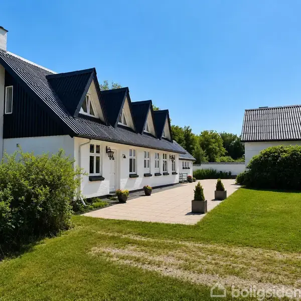 Hvid bygning med sort tag står i solrigt haveområde, omkranset af grønne buske og græs. Flisebelagt gangsti foran huset fører hen imod en garage.