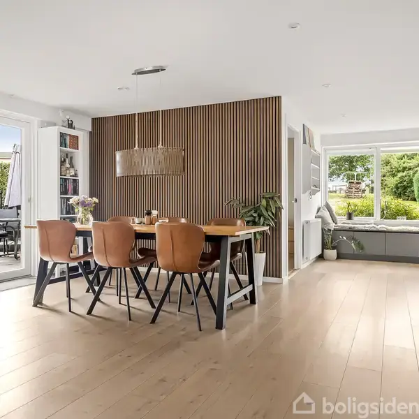 Spisebord med seks stole står i en moderne stue. Stuen har trægulv, en skillevæg med lameller og store vinduer, der lader naturligt lys strømme ind fra haven.