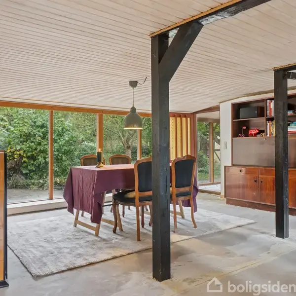 Spisebord med lilla dug er omgivet af otte stole, placeret på et lyst tæppe. Rummet har træhylder til højre og store vinduer, der giver udsigt til grønne omgivelser.