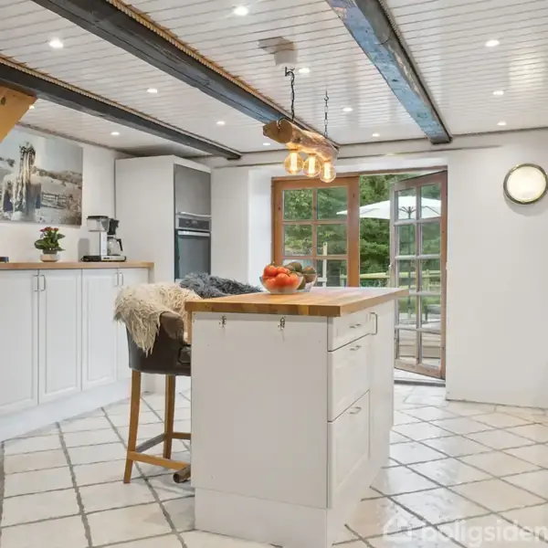 Køkken med hvidt inventar og centrale træbjælker, roligt oplyst af indbyggede spots. Et stort vindue åbner mod grønne omgivelser. Frugt skåle og kaffemaskine står på bordpladerne.