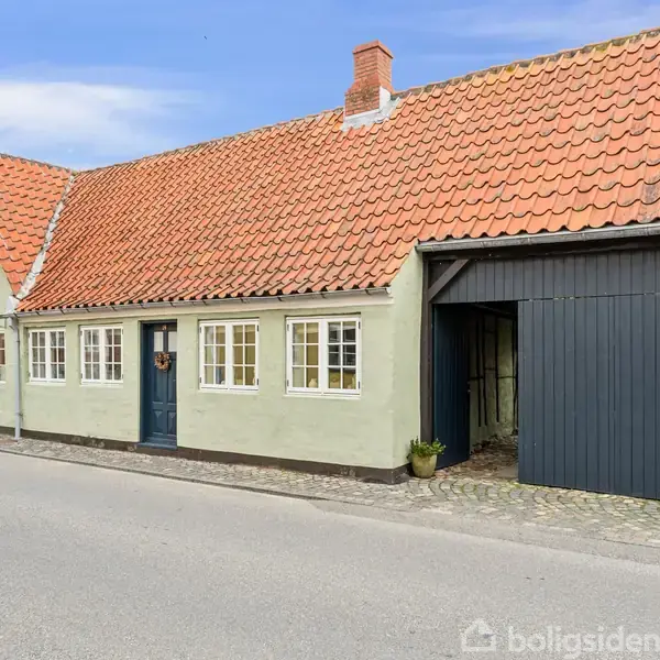 Et klassisk gammeldags hus med røde tegltagsten står på en stille vej. Huset har grønne mure, små sprossede vinduer og en mørkeblå dør ved siden af en åben garage.