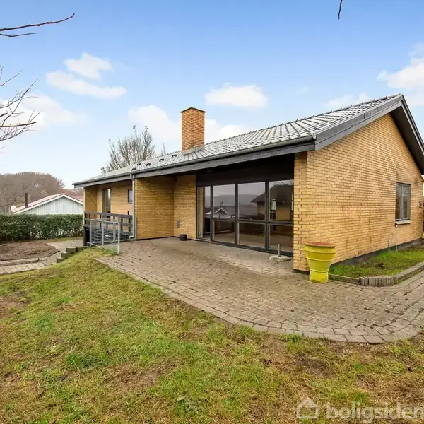 En gulstensvilla beliggende på en græsplæne med terrasse omgivet af fliser. Villaen har store vinduespartier og en skorsten. Træer og buske omkranser området under en blå himmel.