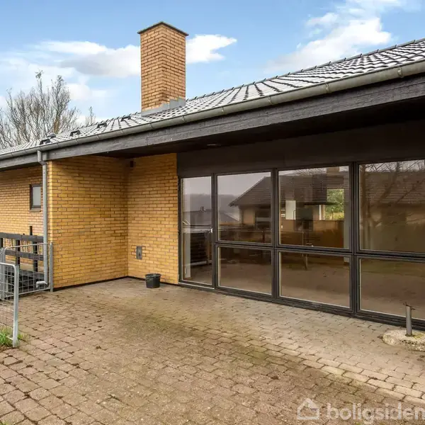 En enkeltstående murstensbygning står stille på en flisebelagt terrasse med et tagudhæng. Store vinduer reflekterer omgivelserne med spredte træer og andre huse i baggrunden.