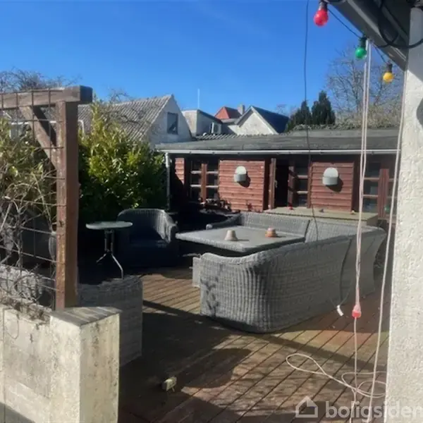 En terrasse med grå sofaer og bord omgivet af planter og en hvid mur. I baggrunden ses et rødt træhus under en klar blå himmel. Farvede lamper hænger over terrassen.