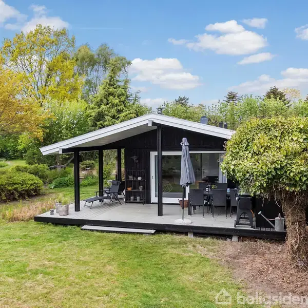 En sortmalet sommerhus med skråtag står i en frodig have. En terrasse med havemøbler og parasol ses foran, og træer omkranser området under en klar, blå himmel.