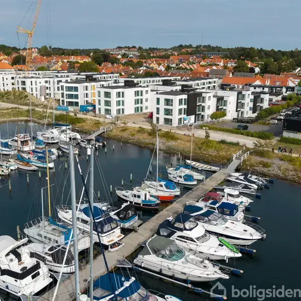 Både ligger fortøjet ved en marina, tæt på en by med moderne og traditionelle bygninger i baggrunden. En kran ses langt væk blandt de rødt tagede huse.