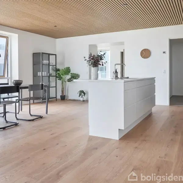 Et moderne køkken med en stor hvid køkkenø. Spisebord med stole ved siden af store vinduer. Rummet har træloft og gulv samt planter og minimalistisk indretning.
