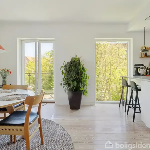 En lys spisestue med et rundt bord, dækket med bordskånere, omgivet af stole. En stor plante står ved et vindue. Køkkenet har barstole ved en disk.