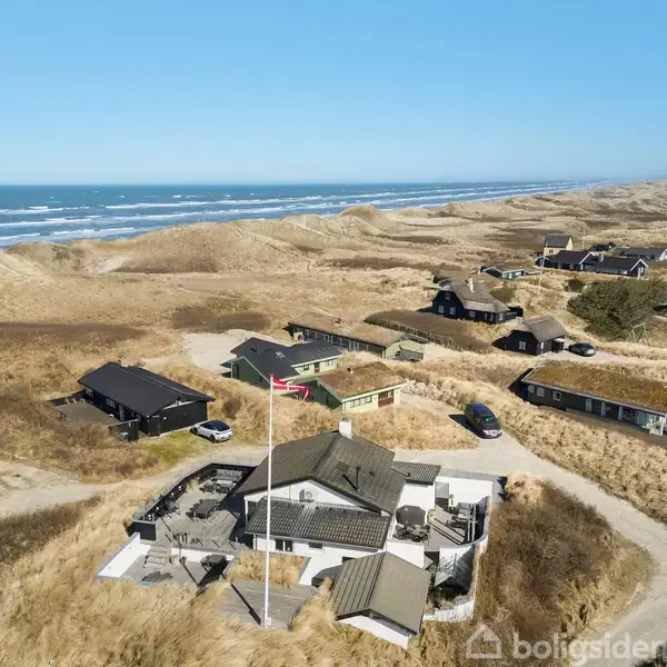 Feriehus med flagstang står stille mellem klitter og græs ved kysten. Havet ses i baggrunden, omkranset af flere spredte sommerhuse.