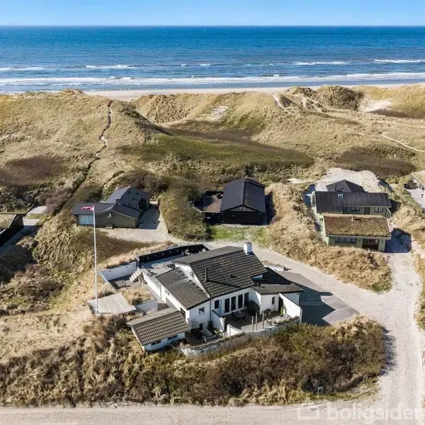 Hus set fra oven med sort tag ligger blandt klitter ved kysten. Omkranset af andre boliger med udsigt over havet og stranden. Ingen synlig tekst.