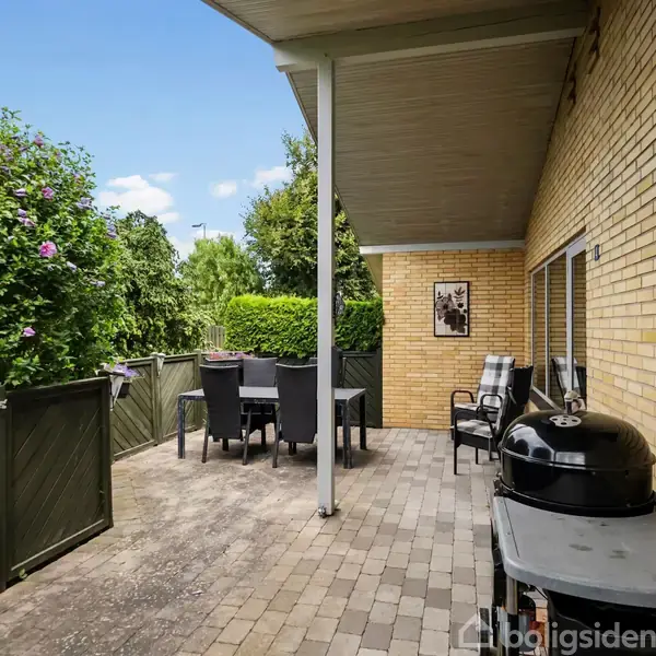 Grill står på overdækket terrasse ved gul murstensvæg, omgivet af havemøbler og grønne buske. Himlen er blå med lette skyer.